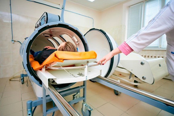 Patient lying inside a traditional medical hyperbaric oxygen chamber while a healthcare professional adjusts the settings.