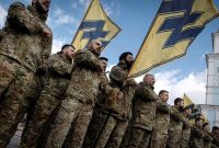 Group of uniformed soldiers standing in formation, holding yellow flags with black symbols, raising their fists during a rally in front of a church with a golden dome.