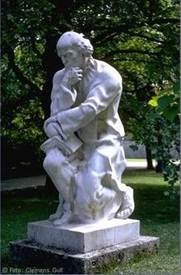 White marble statue of a man sitting and thinking with his hand on his chin, placed in a park surrounded by trees.