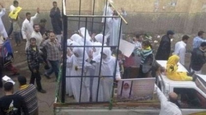 Women dressed in white robes are confined in a cage on the back of a truck during a public demonstration, surrounded by a crowd of onlookers.