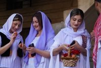 Three Yazidi women in white and lavender traditional clothing holding eggs and a basket, smiling and talking together.