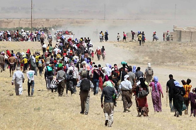 Large group of Yazidis walking through a dusty field, appearing to flee or migrate, carrying belongings.