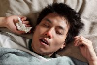 A sick man lies in bed with a red nose, mouth slightly open, and a tissue in his hand, showing symptoms of flu or a severe cold.