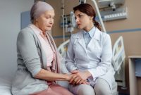 Female doctor sitting beside a woman wearing a headscarf in a hospital room, holding her hands and offering emotional support.