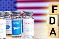 Vials labeled Moderna and Pfizer COVID-19 vaccines standing in front of an American flag, with wooden blocks spelling ‘FDA’ on the right.