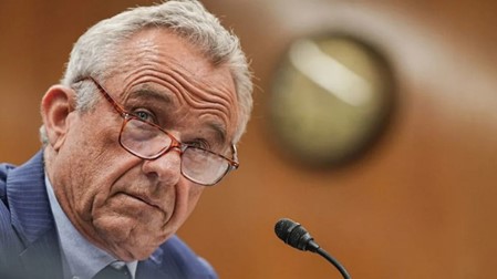 Robert F. Kennedy Jr (RFK Jr.) wearing glasses and a suit speaks into a microphone at a formal hearing, with a blurred circular emblem behind him.