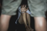 A person sitting on the ground, covering their face and raising one hand as if to shield themselves.