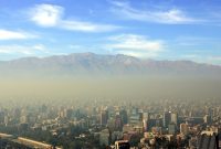 Cityscape with smog covering tall buildings, with mountains visible in the background under a blue sky.