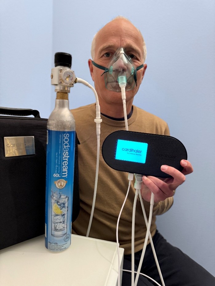 A man sitting indoors wearing a breathing mask connected to tubes, holding a black medical device labeled “Cardihaler” and a CO₂ canister beside him.