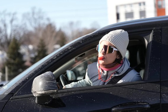 Person sitting in a car wearing winter clothing and sunglasses, speaking through an open driver-side window.