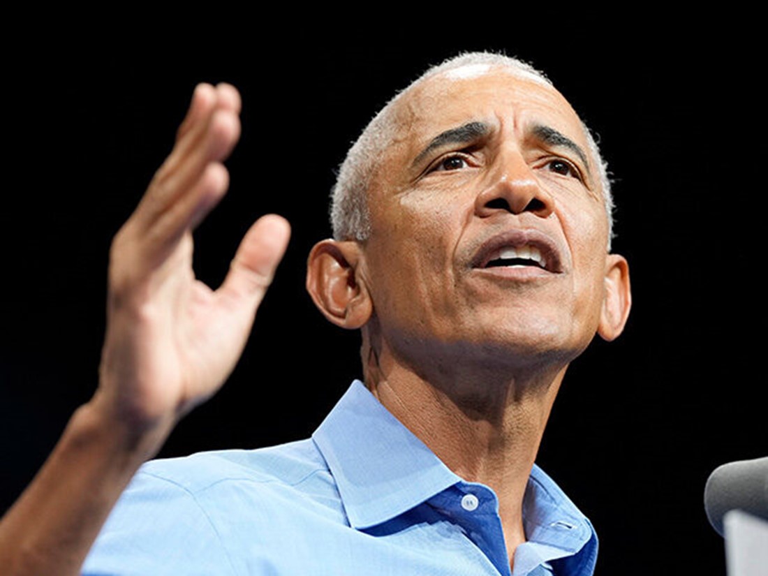 Close-up of Obama speaking at a podium with one hand raised against a dark background.