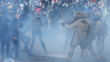Armed soldiers aiming weapons toward a group of masked civilians amid smoke during a tense street confrontation.