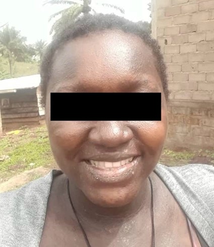 Portrait of a smiling woman outdoors with a black bar covering her eyes, standing near a brick wall and greenery.