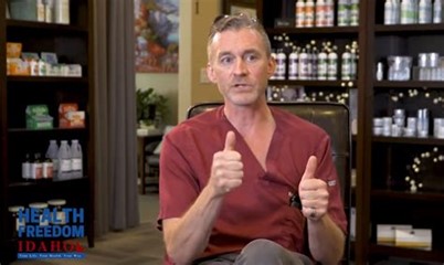 Man seated indoors wearing medical scrubs, gesturing with both hands, with shelves of bottles visible behind him and a small media logo in the corner.