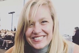 Close-up portrait of a smiling woman wearing a light-colored sweater, photographed outdoors or in soft lighting.