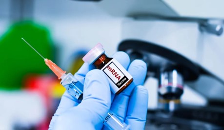Laboratory worker holding a syringe and an mRNA vaccine vial beside a microscope.